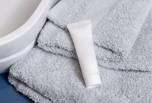 White cosmetic squeeze tube on light grey folded towels near vessel basin in bathroom, close up, brand packaging mockup. Natural handmade cosmetic product