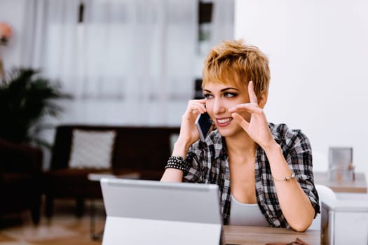 Young woman with short blond hair wearing a plaid shirt is sitting at a desk in her home office, talking on her phone and gesturing with her hand while working on her laptop