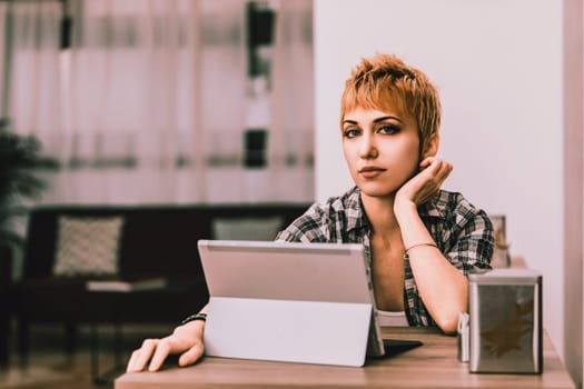 Young woman with short hair, wearing a plaid shirt, sits at a table in a cafe, resting her chin on her hand and looking thoughtfully