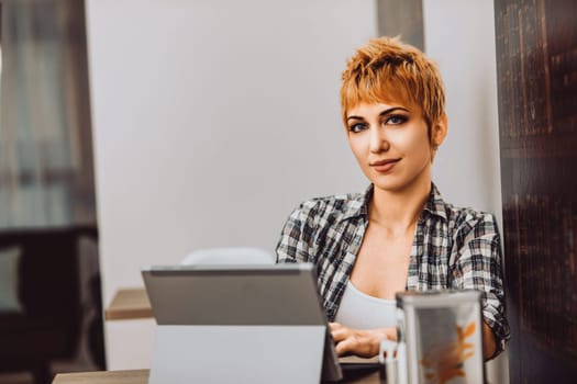 Young woman with short hair is sitting in a cafe, working on her laptop. She is wearing a plaid shirt and smiling slightly