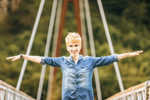 Beautiful young woman with short hair is standing on a bridge with her arms outstretched, smiling cheerfully. She is enjoying the fresh air and the beauty of her surroundings