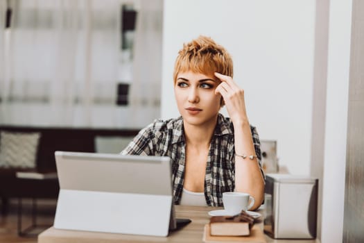 Young freelancer woman with short hair is touching her forehead while thinking about a work project on her laptop in a cafe