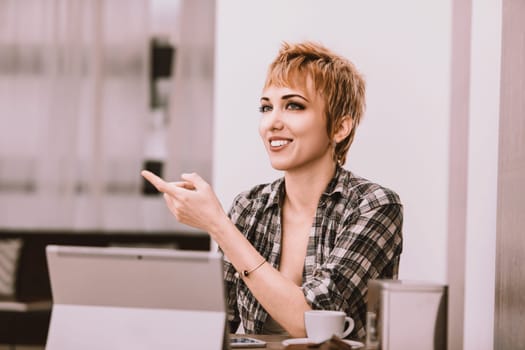 Cheerful female freelancer is sitting in a cafe, enjoying a cup of coffee while working on her laptop. She is pointing with her finger as if explaining a project to a client
