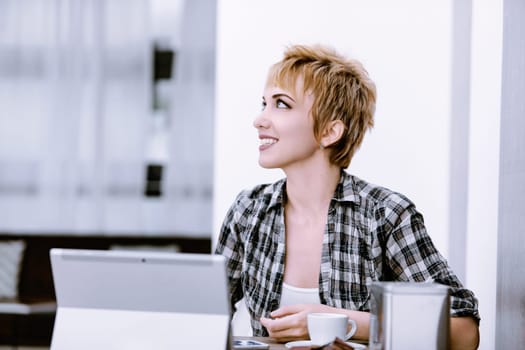 Young woman with short blonde hair is smiling while looking away, sitting at a table with a laptop in front of her and a cup of coffee
