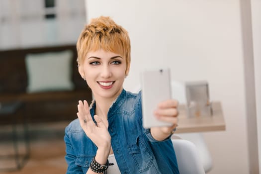 Young woman smiling and waving while taking a selfie with her phone in a modern apartment, exuding a positive vibe in her denim outfit