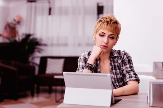 Young woman with short blonde hair looks concerned working from home on tablet, focusing on a problem