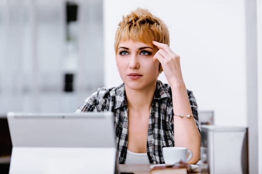 Young woman with short blonde hair is sitting at a table, touching her head and thinking. She is wearing a plaid shirt and has a cup of coffee in front of her