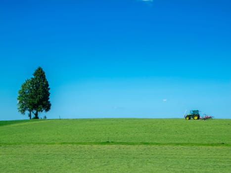 Landscape view of a freshly mown pasture with a single tree and tractor with mowing trailer under a blue sky in Bavaria.