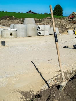 View of a construction site in the Bavarian Allgäu with concrete pipe elements for the construction of sewage canals in the background and a vertical shovel in the foreground in summer sunshine.