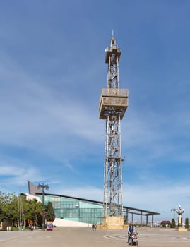 Baku, Azerbaijan - May 10, 2024: Metal parachute tower looms over a paved area with people walking by. Modern business center building is visible in the background