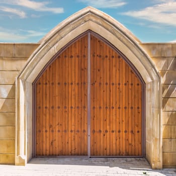 A pair of wooden doors with decorative metal accents are set within a stone archway. The doors are closed, and the sunlight casts shadows on the stone walls surrounding the entrance