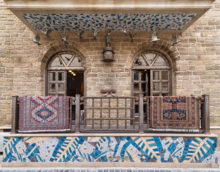 A view of a building in Old Town Baku, Azerbaijan. The balcony has a tiled roof and iron railings. Two decorative rugs hang on the railing, adding a splash of color to the scene