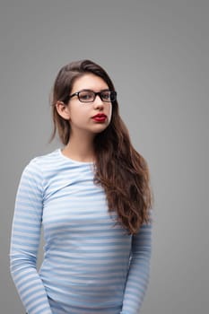 Young woman with long brown hair wearing glasses and red lipstick is posing on a grey background, her expression serious and contemplative