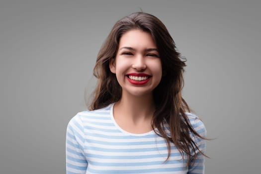 Young woman with long brown hair and a striped shirt smiles playfully with red lipstick in a grey studio, radiating happiness and confidence