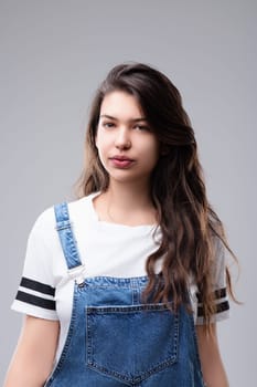 Young woman with long brown hair is standing against a grey background, wearing a white t shirt and denim overalls. Her neutral expression conveys a sense of confidence and self assurance