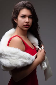 Stunning young latin woman exudes elegance in a red dress with a white fur stole, radiating confidence and allure against a soft grey backdrop