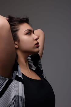 Young woman is posing in a studio, holding her hair and looking away from the camera. She is wearing a black and white cardigan and a black top