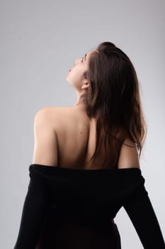 Young woman with long brown hair wearing an elegant black dress is posing in studio, she's holding her hair and looking up