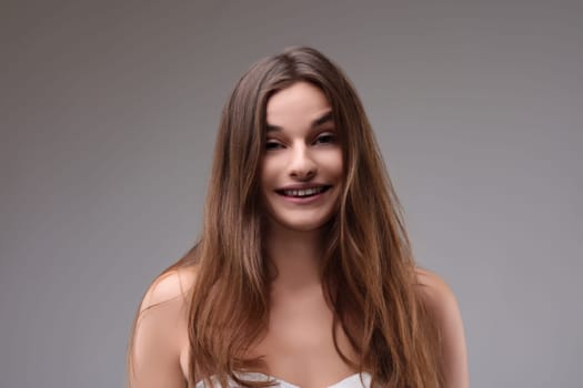 Portrait of a beautiful young woman with long brown hair smiling on a grey background, expressing happiness and positive emotions