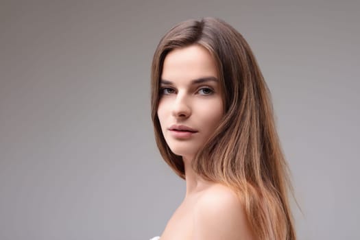 Beautiful young woman poses serenely with long brown hair against a neutral grey background, exuding confidence and calm