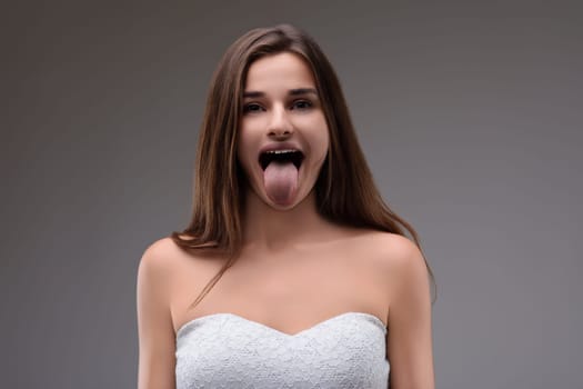 Carefree young woman with long brown hair playfully sticks out her tongue against a gray backdrop, wearing a chic white dress, exuding joy and humor. Perfect for a fun and lighthearted vibe