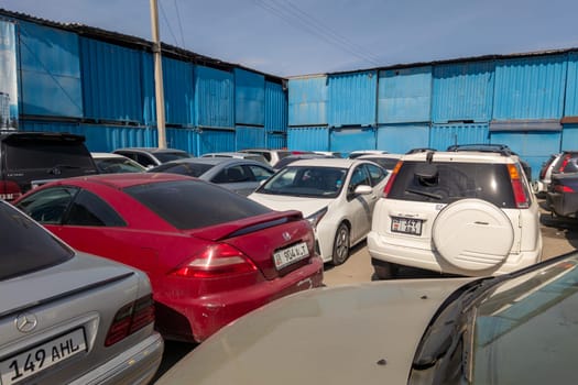 many cars densely parked near blue trading containers during sunny day at Dordoi Bazaar in Bishkek, Kyrgyzstan - March 20, 2023