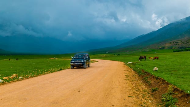 old SUV driving along dry dirt road near grazing horses before thunderstorm in Semenovskoye gorge, Kyrgyzstan at July 5, 2023