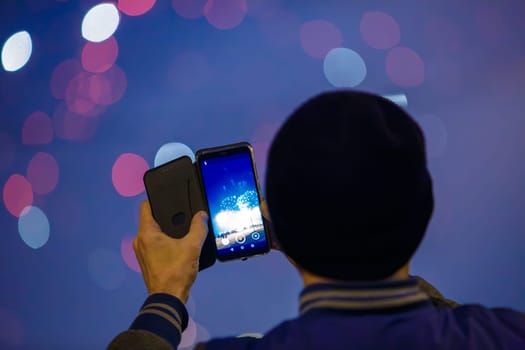 unrecognizable man with black hat photographing fireworks at night with cellphone, closeup with selective focus, rear view