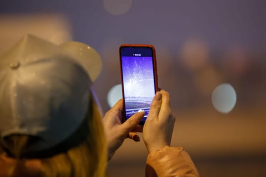 unrecognizable blonde woman with silver cap photographing fireworks at night with cellphone, closeup with selective focus, rear view