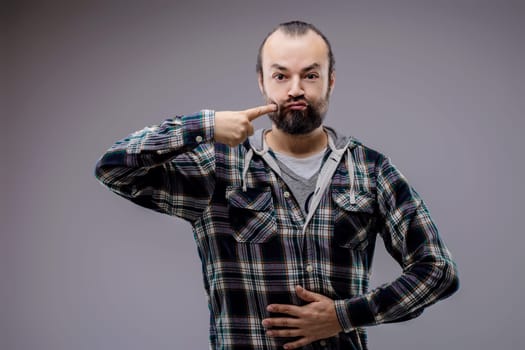 Bearded man playfully touching cheek, making funny face and puffing cheeks, exuding humor and amusement in gray studio setting