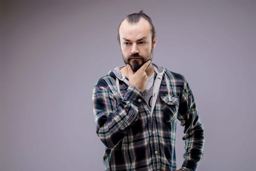 Bearded man wearing a plaid shirt is holding his chin with a thoughtful expression, trying to make a decision or find a solution