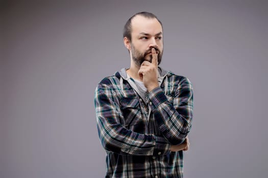 Bearded man wearing a plaid shirt is holding his chin with his finger, deep in thought and looking away from the camera