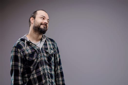 Young man in a plaid shirt looks up with a hopeful smile, lost in thought against a grey backdrop. Portrait of a positive, casual millennial