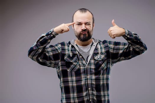 Bearded man pointing a finger to his temple and showing thumbs up with a grimace on his face, expressing a challenging concept