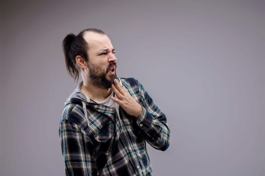 Casual man with beard and ponytail wearing a plaid shirt, touching his face with a disgusted expression on a neutral background
