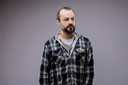 Bearded man in a plaid shirt and hoodie looks thoughtful and worried against a gray background, embodying masculinity and solitude with a stylish appearance