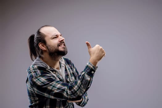 Bearded man in a plaid shirt gives a thumbs up with a funny expression. Symbolizing agreement and satisfaction. He is recommending or endorsing something in a studio shot against a grey background