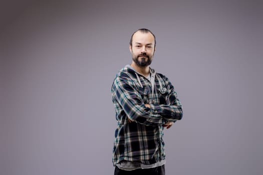 Confident bearded man posing with folded arms in stylish plaid shirt, looking content and happy in studio setting with neutral gray background