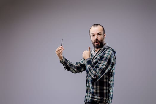 Surprised man takes a selfie with a smartphone, showing happiness and wonder. He gives a thumbs up in a studio with a gray background, wearing a casual checkered shirt