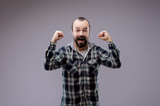 Man with a beard is expressing positive feelings by raising his fists in the air and opening his mouth wide while standing in front of a grey background