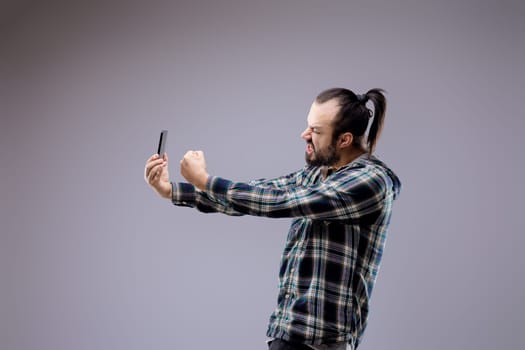 Bearded man with a ponytail shows intense emotions while using a smartphone, reflecting stress and overwhelm from technology's impact on mental health in younger generations