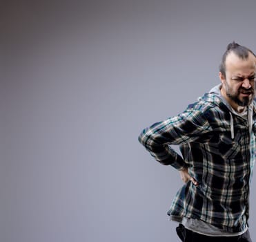 Man in his 30s grimacing in pain, holding his lower back due to backache, showcasing negative emotions in a studio shot against a gray background