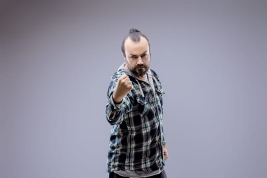 Aggressive man with a beard wearing a plaid shirt is throwing a punch with a threatening facial expression on a gray background