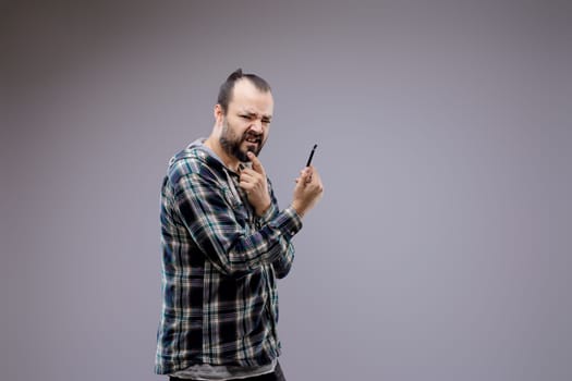 Man with a beard is holding a pen and looking suspicious while making a face. He is standing in front of a grey background