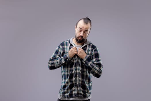 Studio portrait of a bearded man with his hair tied back, wearing a plaid shirt over a hooded sweatshirt, buttoning his shirt with a thoughtful expression