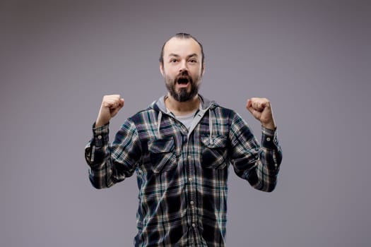 Bearded man celebrates with raised fists, showing pure joy and excitement, radiating success and achievement in a studio shot against a gray background