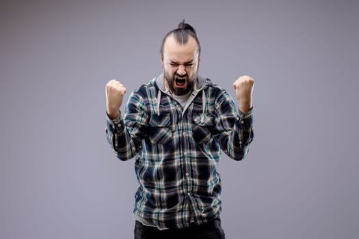 Young man is posing in a studio, wearing casual clothing and raising his fists in the air while shouting. He is experiencing powerful feelings of happiness and victory