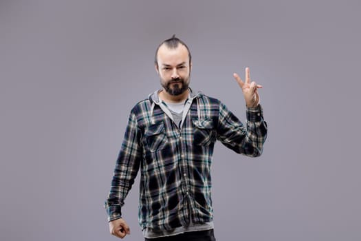 Bearded man in a plaid shirt energetically gesturing rock and roll against a gray background, exuding cool confidence and rebellion as he gazes at the camera