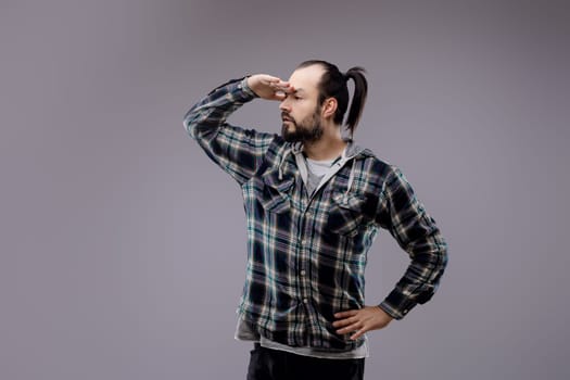 Man with a beard and ponytail is standing with one hand on his hip and the other shielding his eyes as he looks intently into the distance