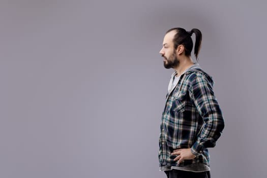 Bearded man in a plaid shirt, standing against a gray background, lost in deep thought, exuding masculinity and solitude with a trendy hipster style